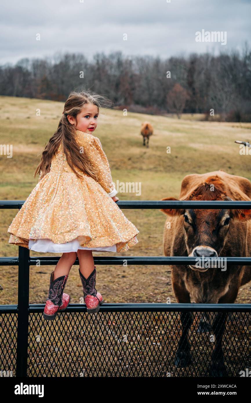 Ragazza in abito da paillettes oro sul recinto con mucche sul campo Foto Stock