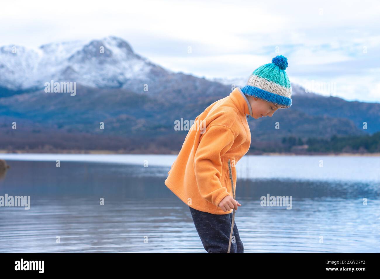 Ragazzo caucasico in un cappello di lana che gioca nella natura in un paesaggio di un lago e montagne innevate. Concetto di bambini sani Foto Stock