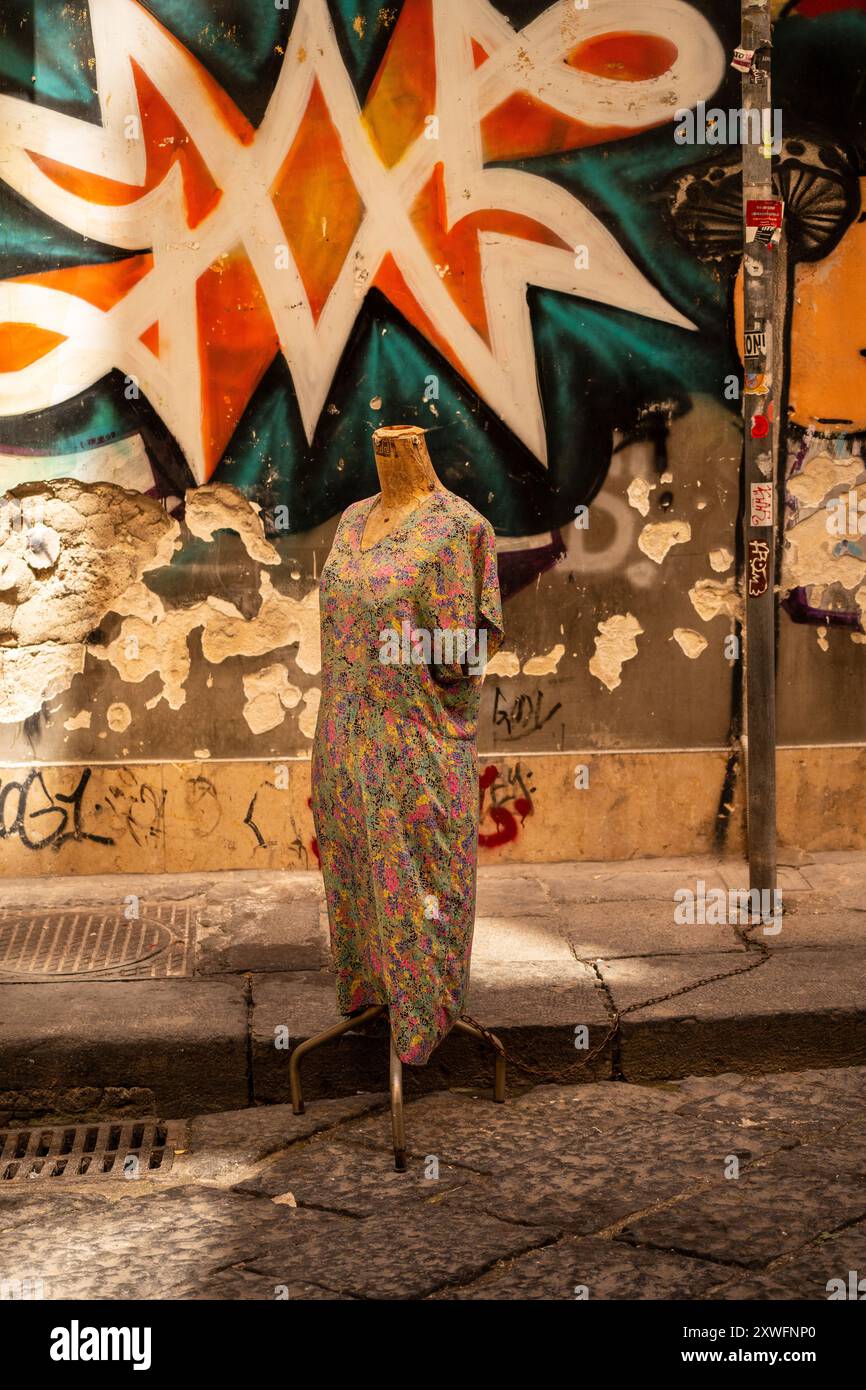 Napoli, Italia - 22 giugno 2024: Un manichino vintage vestito con un abito colorato si erge su una parete ricoperta di graffiti, che fonde moda e stile urbano Foto Stock
