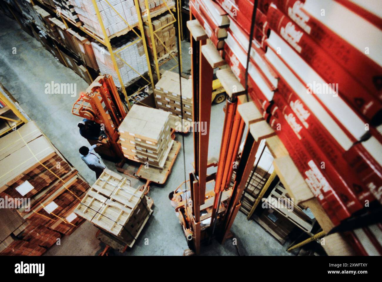 Due operatori manovrano i carrelli elevatori intorno ai pallet impilati in un magazzino affollato pieno di scatole e scaffali. Foto Stock
