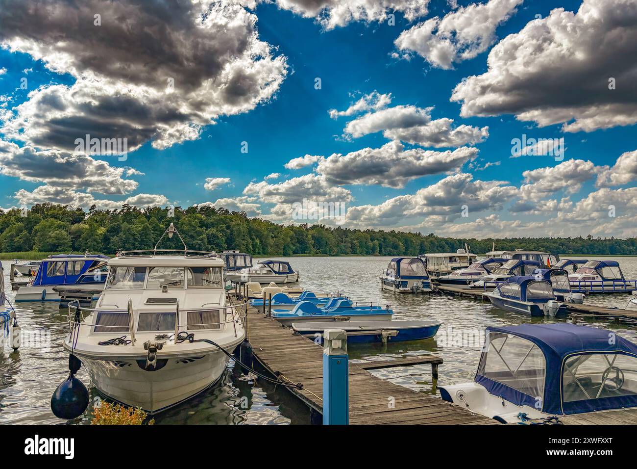 Un gruppo di barche è ormeggiato su Un molo sulla riva di Un lago. Il cielo è nuvoloso e la superficie dell'acqua è calma. Gli alberi possono essere visti nel Backgro Foto Stock