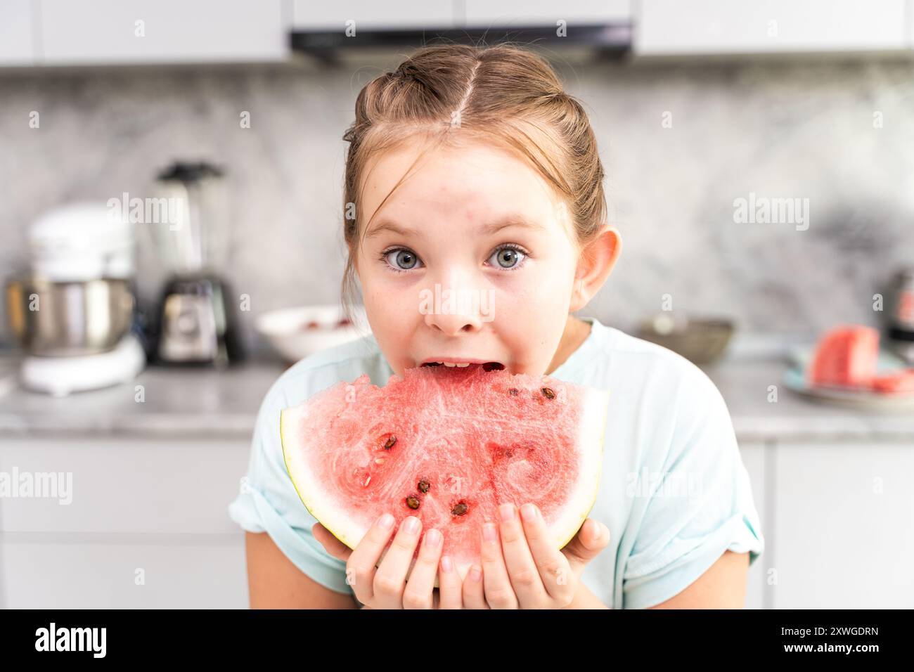 Una bambina morde un pezzo di anguria mentre si trova in cucina a casa con una morbida t-shirt turchese. Foto di alta qualità Foto Stock