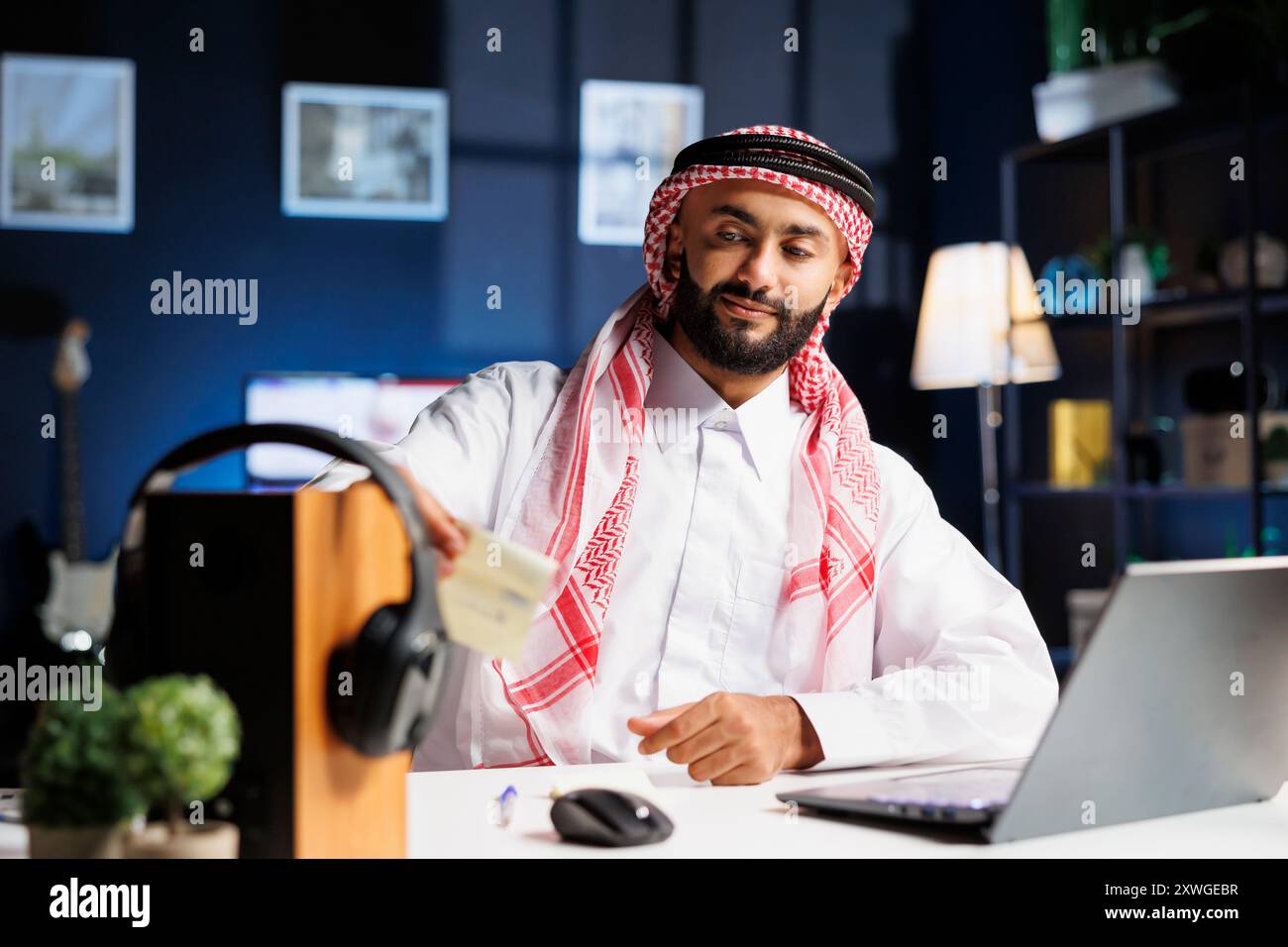 Bell'uomo d'affari che indossa un abito islamico, seduto nel suo ufficio a lavorare sul suo portatile e a scrivere appunti. Giovane musulmano che mette la sua nota adesiva su una tavola di legno con le cuffie. Foto Stock