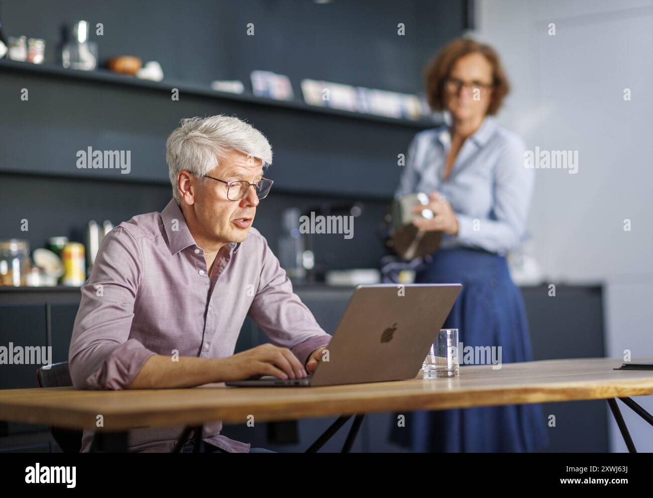 Symbolfoto zum Thema Arbeitsteilung bei Paaren im Haushalt. Ein Mann sitzt in einer Kueche am laptop waehrend eine Frau im Hintergrund Geschirr spuelt Foto Stock