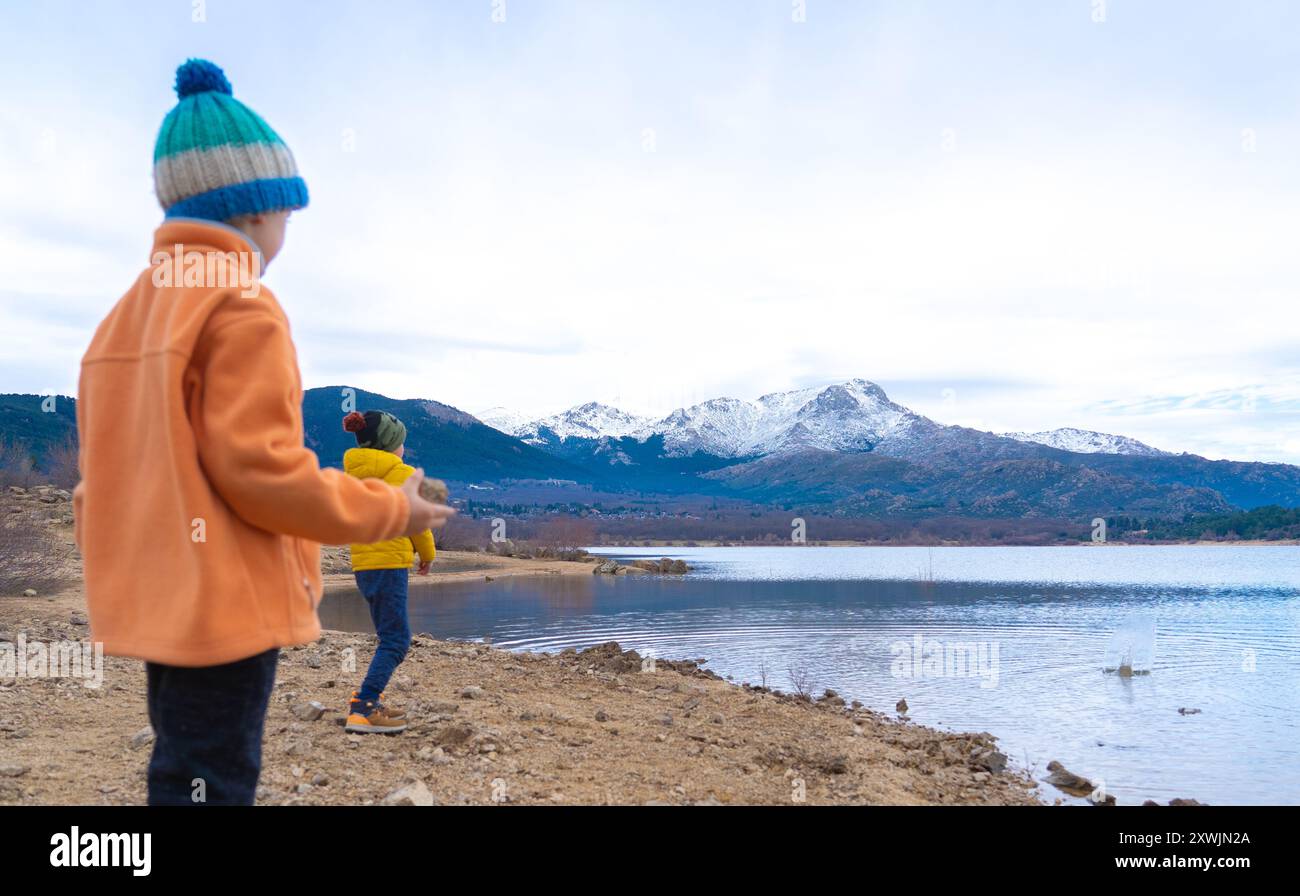 Due bambini che giocano lanciando pietre in un lago con un paesaggio di montagne innevate sullo sfondo in una giornata fredda Foto Stock