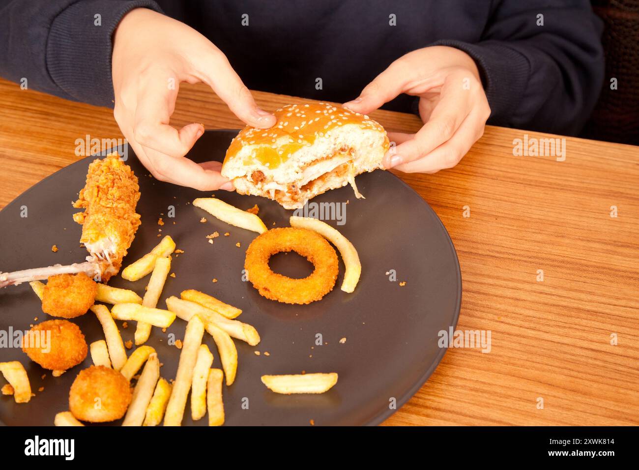 Un uomo caucasico mangia hamburger a metà pasto, patatine, anelli di cipolla e pezzi di pollo a mano da un piatto grigio su un tavolo di legno Foto Stock