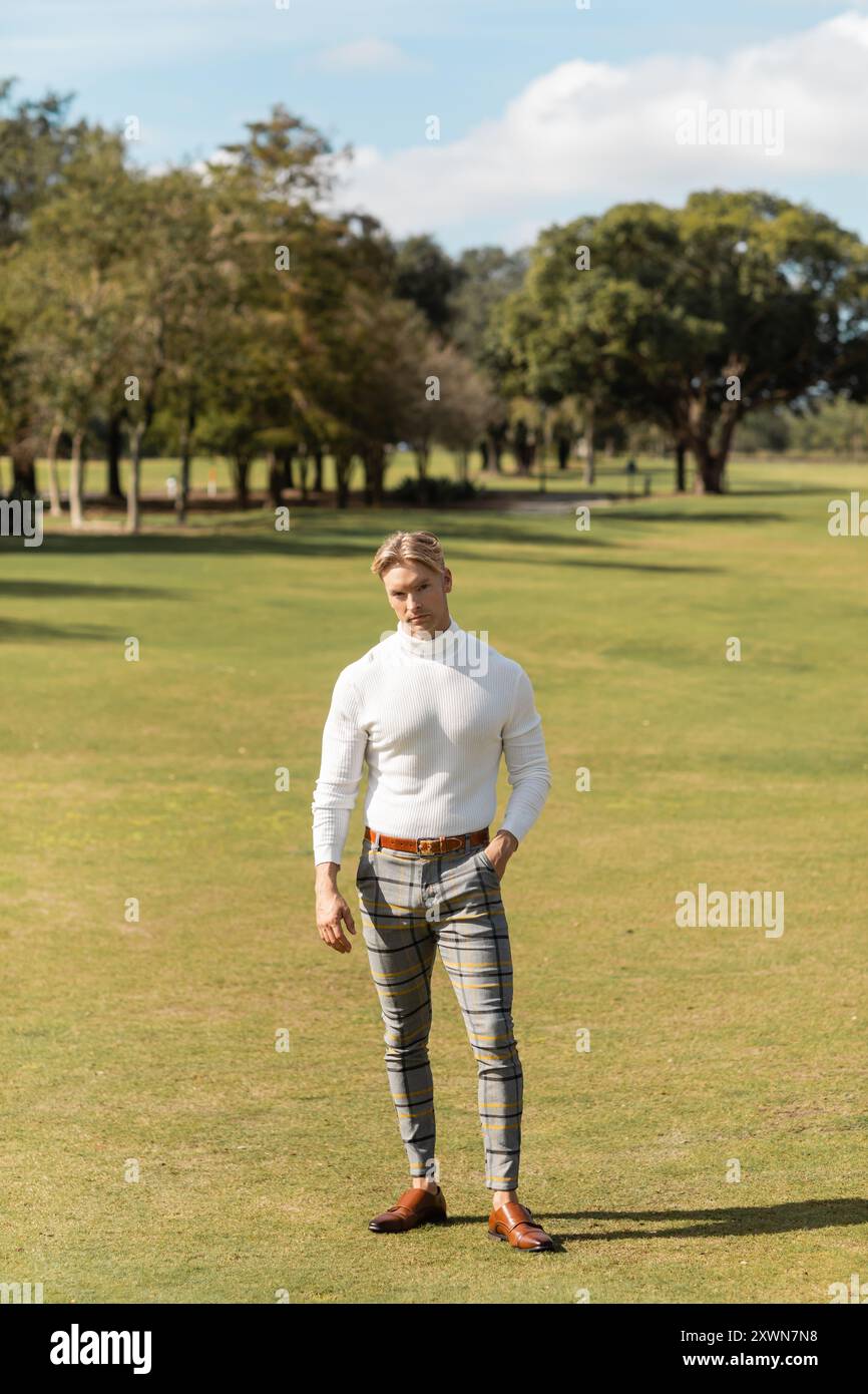 Un uomo biondo con dolcevita bianco e pantaloni fantasia attraversa un lussureggiante parco verde di Miami. Foto Stock