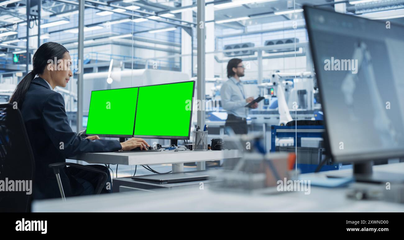 Tecnico robotico donna che lavora su un computer desktop con schermo verde Chromakey Mock Up display in un ufficio di fabbrica. Strumento di ricerca e sviluppo tecnologico con linea di assemblaggio autonoma Foto Stock