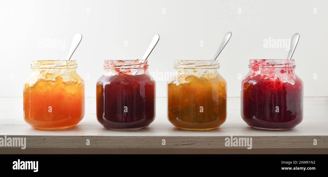 Raccolta di vasetti di marmellata aperti di vario tipo con cucchiaio su tavolo in legno bianco e sfondo bianco isolato. Vista frontale. Foto Stock