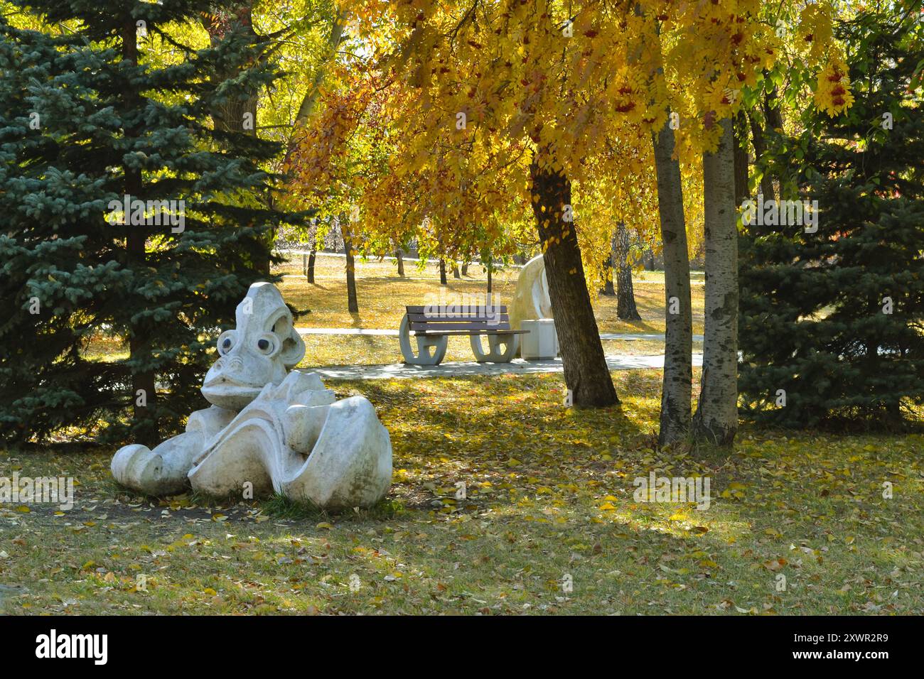 Una scultura di una strana creatura e una panchina in un parco autunnale soleggiato Foto Stock