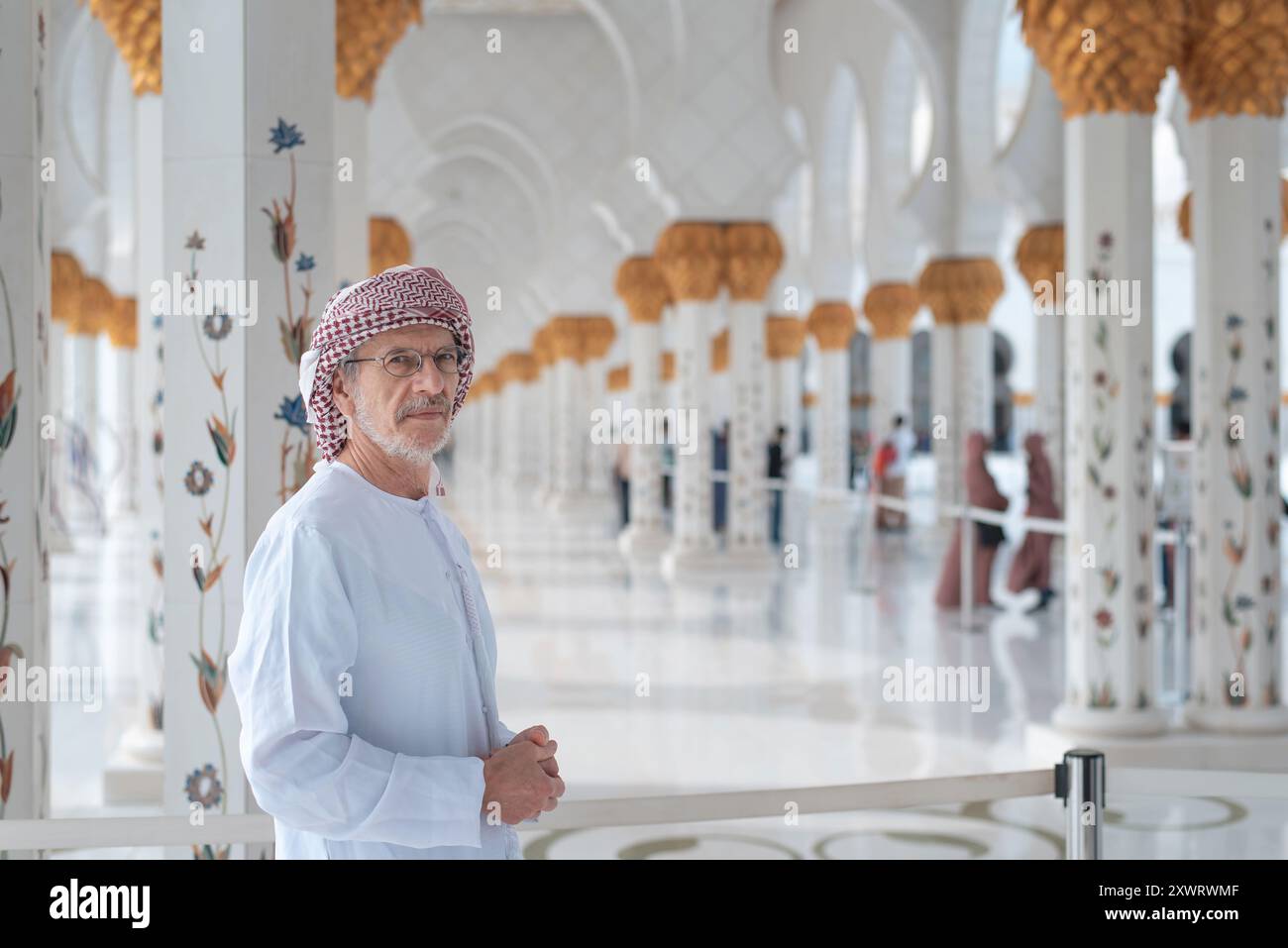 Un uomo vestito con abiti tradizionali si trova all'interno della grande moschea Sheikh Zayed ad Abu Dhabi, circondata da colonne ornate splendidamente decorate con oro Foto Stock
