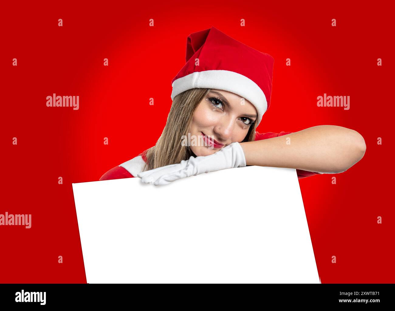 Il giovane Babbo Natale latina sorride guardando la macchina fotografica con un poster vuoto per inserire pubblicità o messaggi su sfondo rosso Foto Stock