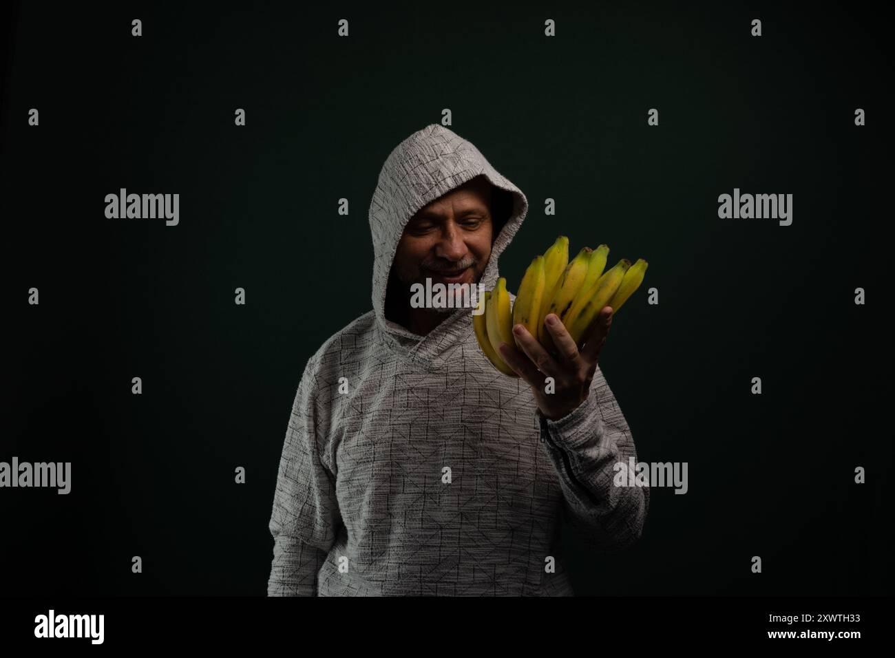 Uomo misterioso che indossa una felpa con cappuccio che tiene un mucchio di banane gialle. Isolato su sfondo scuro. Foto Stock