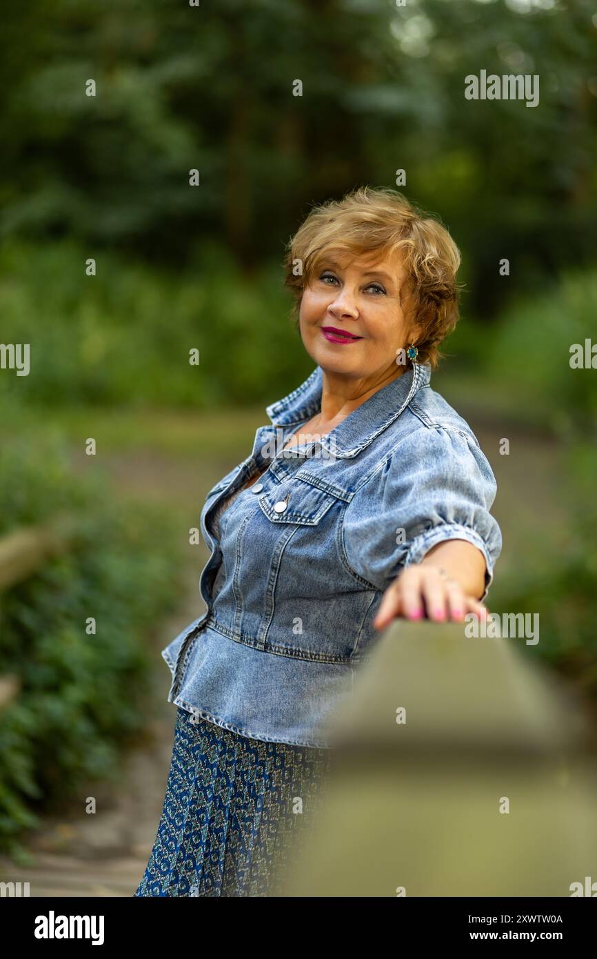 Una donna si trova vicino a una ringhiera di legno, vestita con una giacca in denim e un abito a motivi geometrici, circondata da una vivace vegetazione. Foto Stock