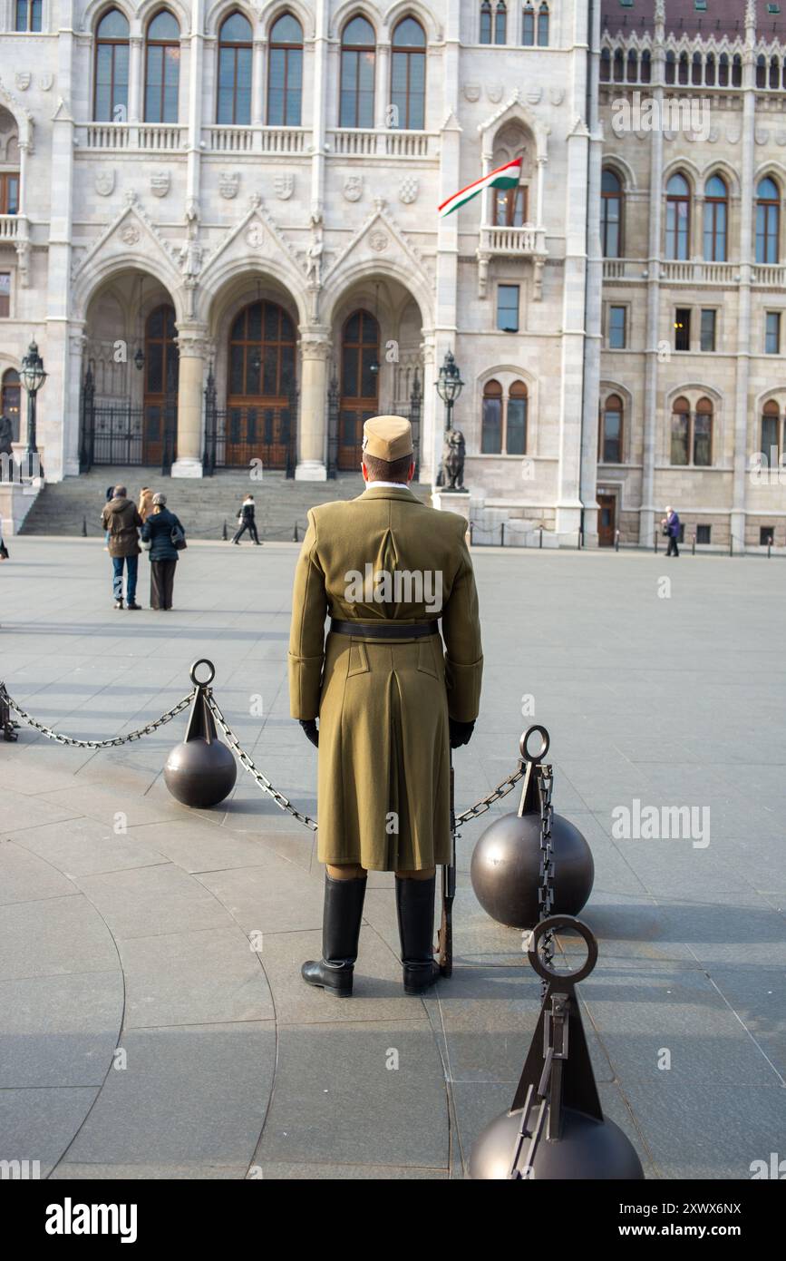 Un soldato si erge in uniforme tradizionale, di fronte al maestoso palazzo del Parlamento ungherese a Budapest. La scena cattura un mix di orgoglio nazionale e grandezza architettonica. La luce del sole mette in risalto gli intricati dettagli della struttura storica. Foto Stock