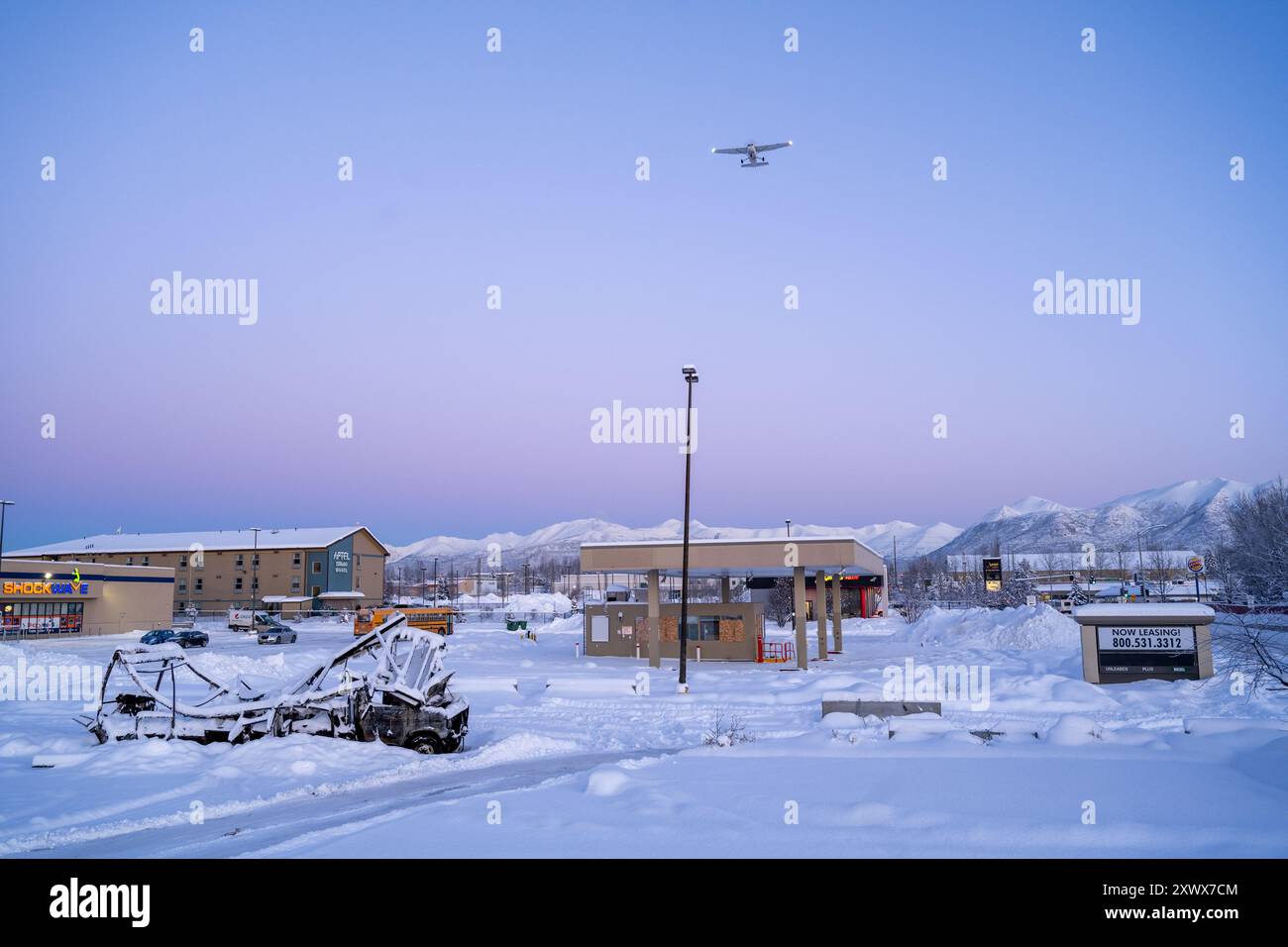 Una vista panoramica invernale con edifici innevati e un aereo che vola vicino all'aeroporto Merrill Field con maestose montagne sullo sfondo, Anchorage, Alaska. Questa immagine evoca temi di tranquillità e resilienza. Foto Stock