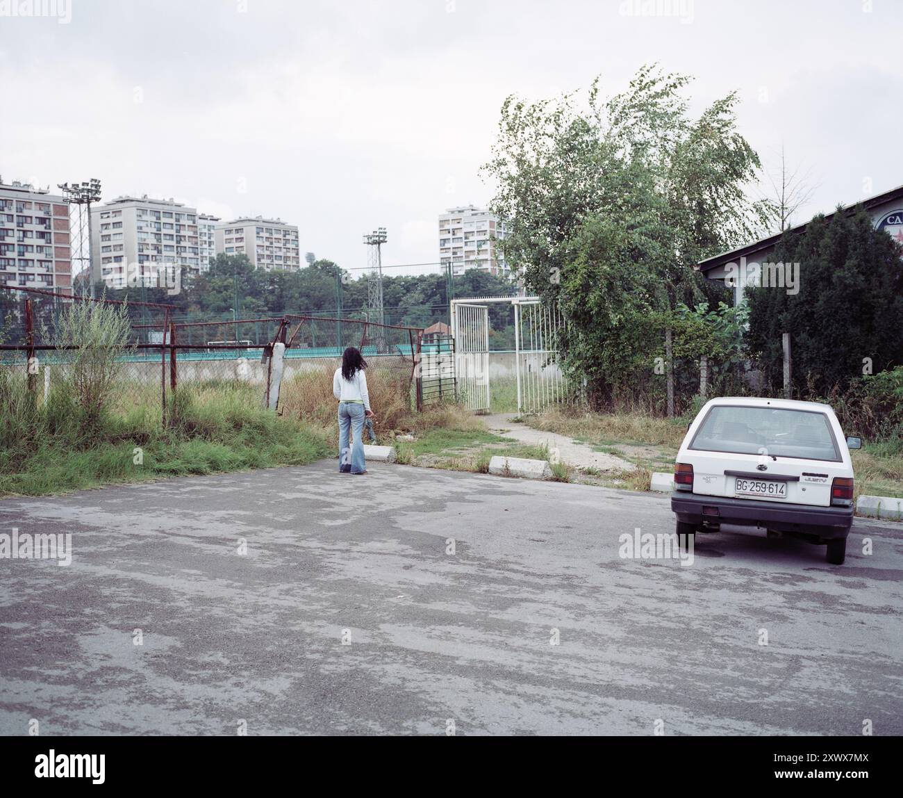 Una donna che passeggia vicino a un'area industriale, simboleggia la solitudine in mezzo allo sviluppo urbano. La scena presenta alti edifici di appartamenti e un campo d'erba recintato, che enfatizza il contrasto tra la natura e la vita cittadina. Belgrado, 2002 Foto Stock