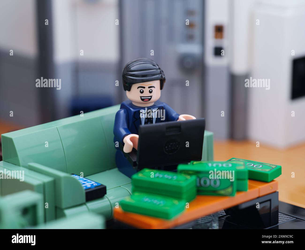 Tambov, Federazione Russa - 18 agosto 2024 Una minifigura d'affari Lego seduta su un divano e che lavora su un computer portatile dietro un tavolo con i soldi. Foto Stock