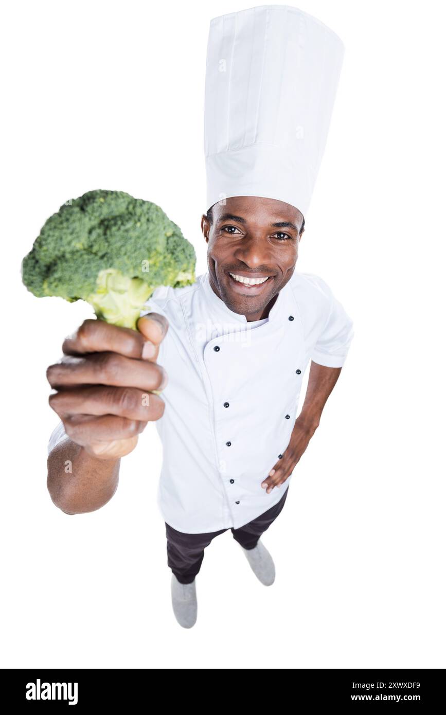 Mangiare sano! Vista dall'alto del giovane e allegro chef africano in uniforme bianca che si estende sui broccoli mentre si trova su sfondo bianco Foto Stock