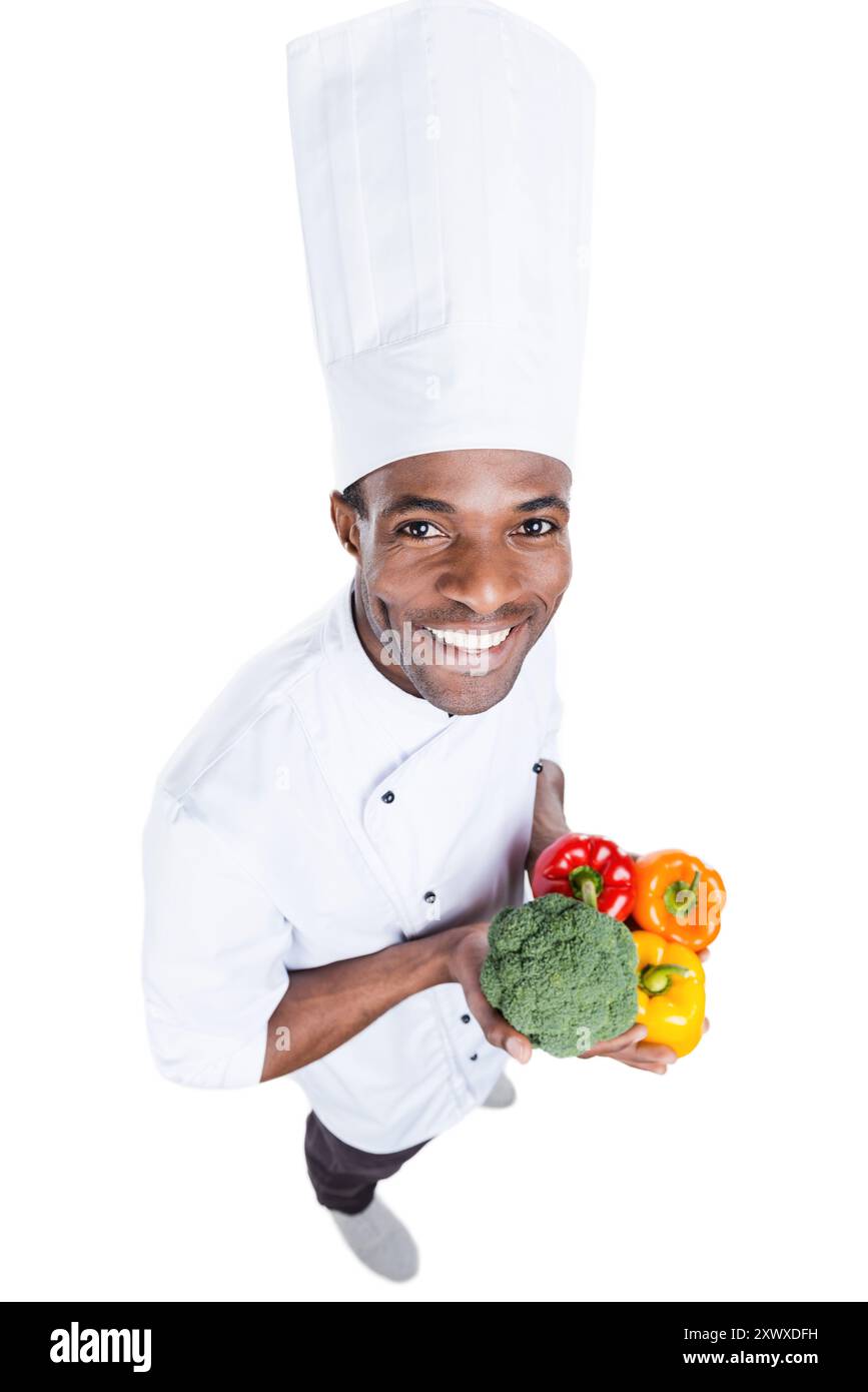 Il cibo sano è nelle sue mani. Vista dall'alto del giovane chef africano in uniforme bianca che regala verdure colorate mentre si trova sullo sfondo bianco Foto Stock