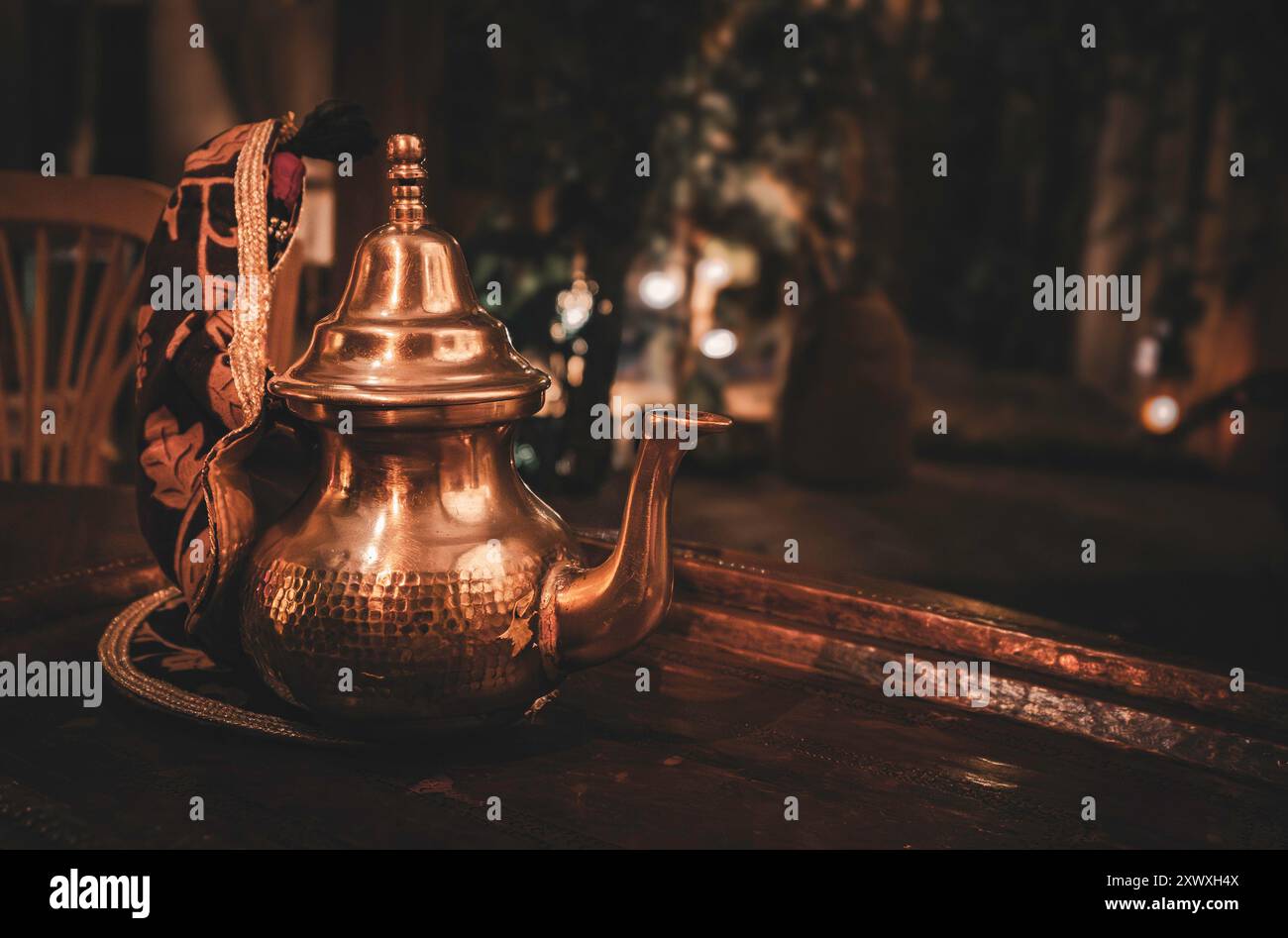 Una teiera in metallo marocchino splendidamente lavorata con disegni intricati, appoggiata su un rustico tavolo in legno. Lo sfondo è leggermente illuminato, creando un ambiente caldo Foto Stock