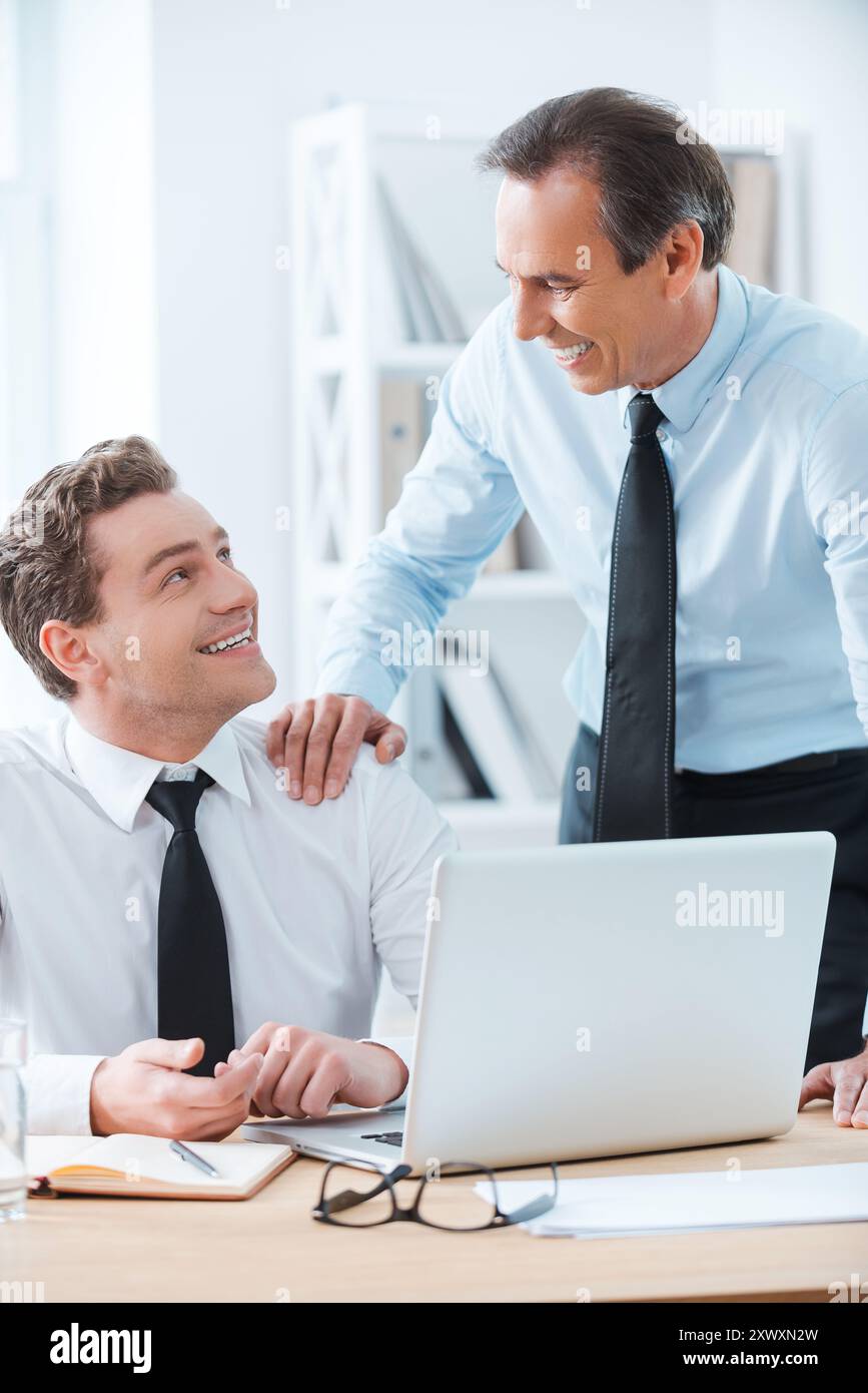 Condividere una buona esperienza. Allegro uomo d'affari seduto al suo posto di lavoro mentre il suo capo si tocca la spalla e si trova vicino Foto Stock