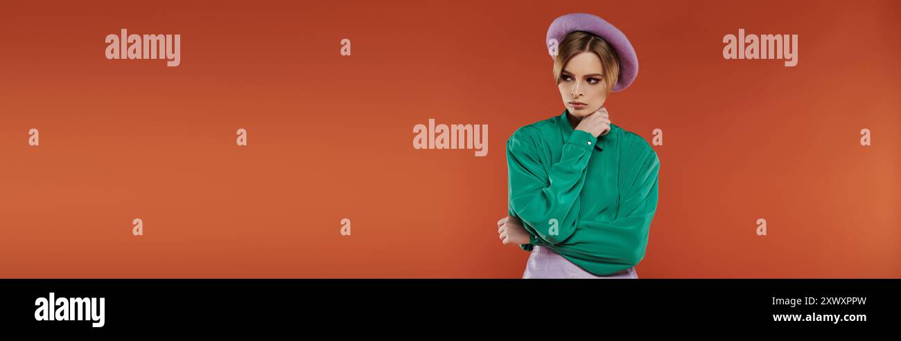 Una donna che indossa una camicia verde e un berretto viola poggia su uno sfondo arancione brillante. Foto Stock