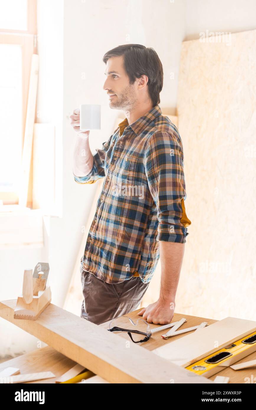 Un sorso di immaginazione. Vista laterale di un giovane falegname che tiene in mano la tazza di caffè mentre si appoggia al tavolo di legno con diversi utensili da lavoro Foto Stock