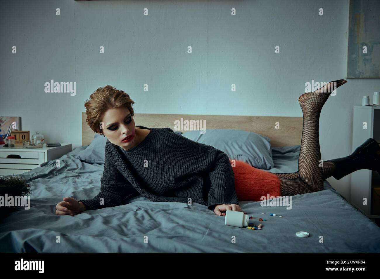 Una donna che indossa calze a rete nere e una gonna rossa si stende su un letto, tenendo in mano una bottiglia di pillola. Foto Stock