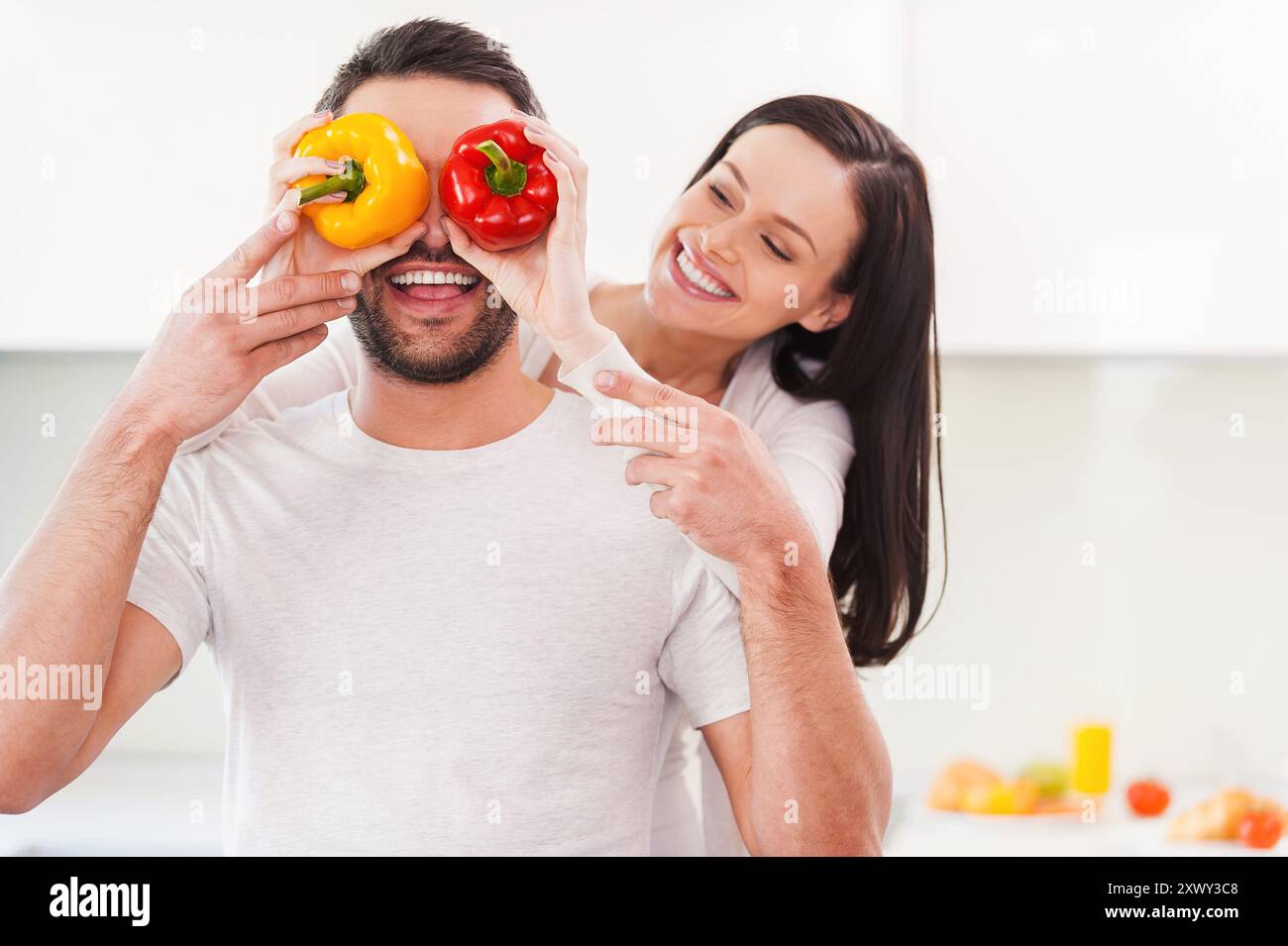 Coppia giocosa. Bella giovane donna che copre gli occhi del suo ragazzo con peperoni colorati e sorride mentre entrambi sono in piedi in cucina Foto Stock Coppia giocosa. Bella giovane donna che copre gli occhi del suo ragazzo con peperoni colorati e sorride mentre entrambi sono in piedi in cucina Foto Stock