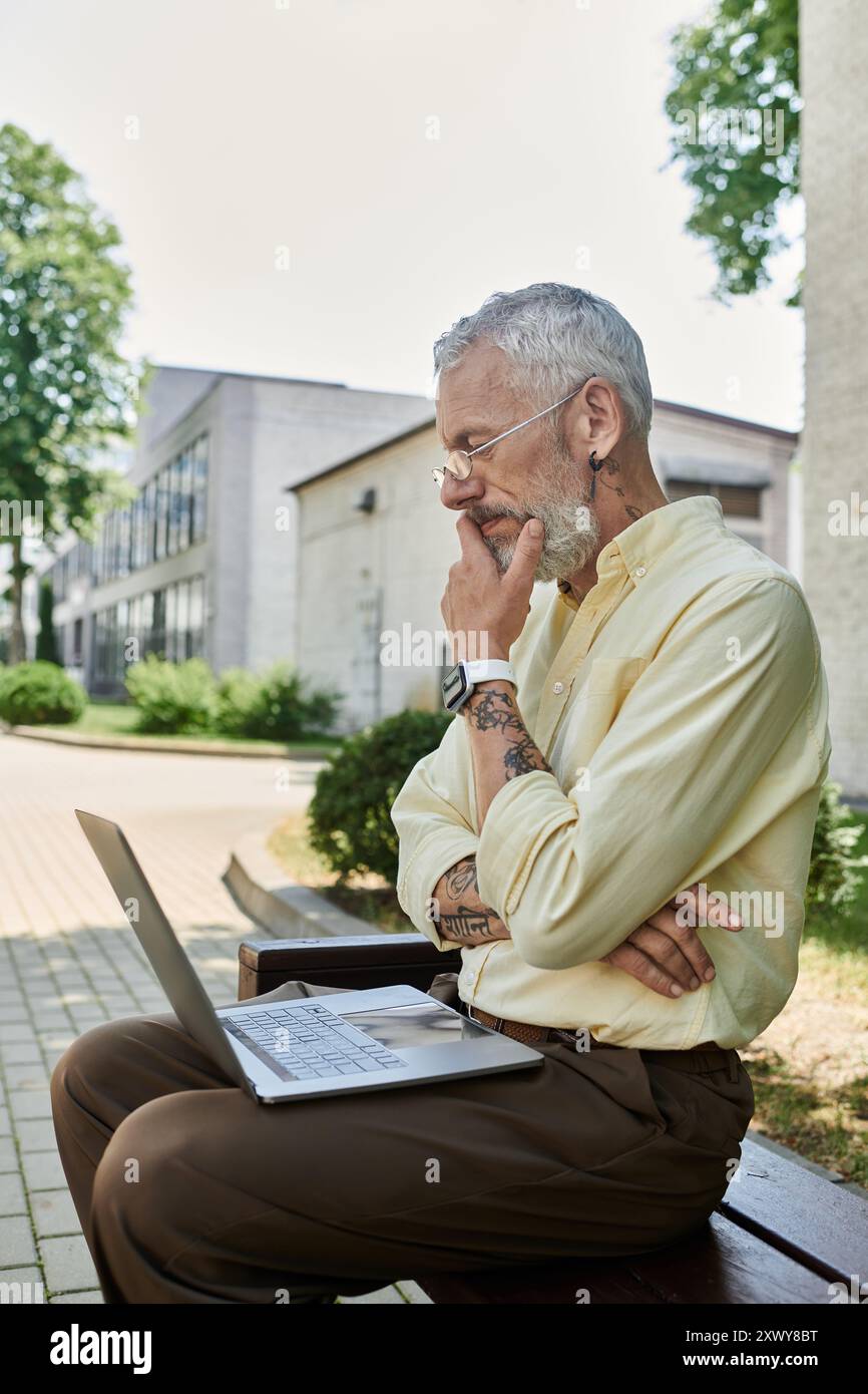 Un uomo maturo con la barba siede su una panchina fuori da un edificio moderno, usando il suo computer portatile e con un pensiero profondo. Foto Stock
