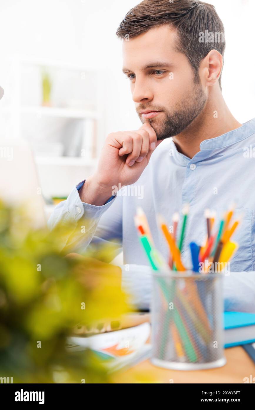 Concentrati sul lavoro. Bel giovane in camicia che lavora su un computer portatile e tiene per mano il mento mentre si siede sul posto di lavoro Foto Stock