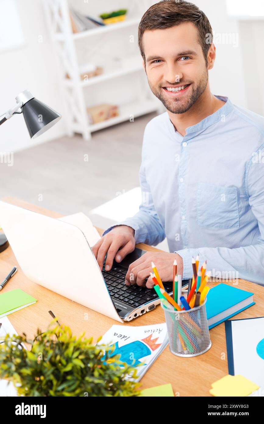 Mi piace il lavoro che fai. Bel giovane in camicia che lavora su un laptop e sorride alla macchina fotografica mentre si siede sul suo posto di lavoro Foto Stock