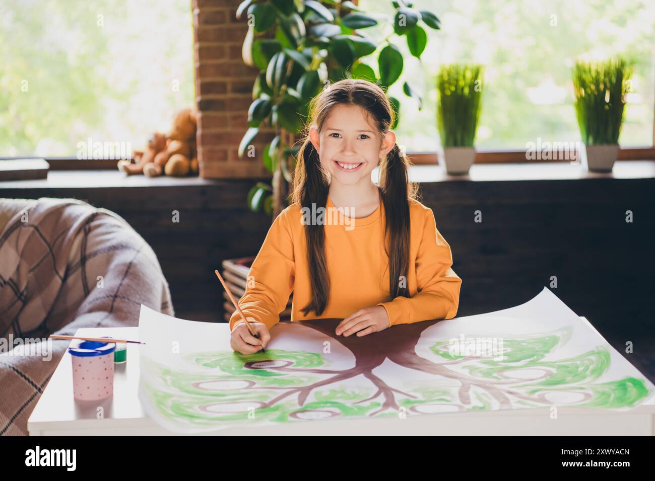 Foto di una bella bambina seduto a tavola disegnando i compiti dell'albero genealogico indossare abiti arancioni interni moderni piatti interni Foto Stock