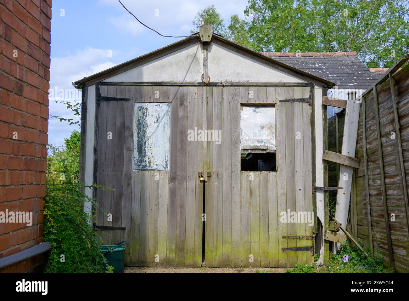 Una capanna in legno vintage, garage, con esterno intempestivo, circondato da vegetazione e recinzioni, la luce naturale brillante mette in risalto il suo fascino rustico Foto Stock