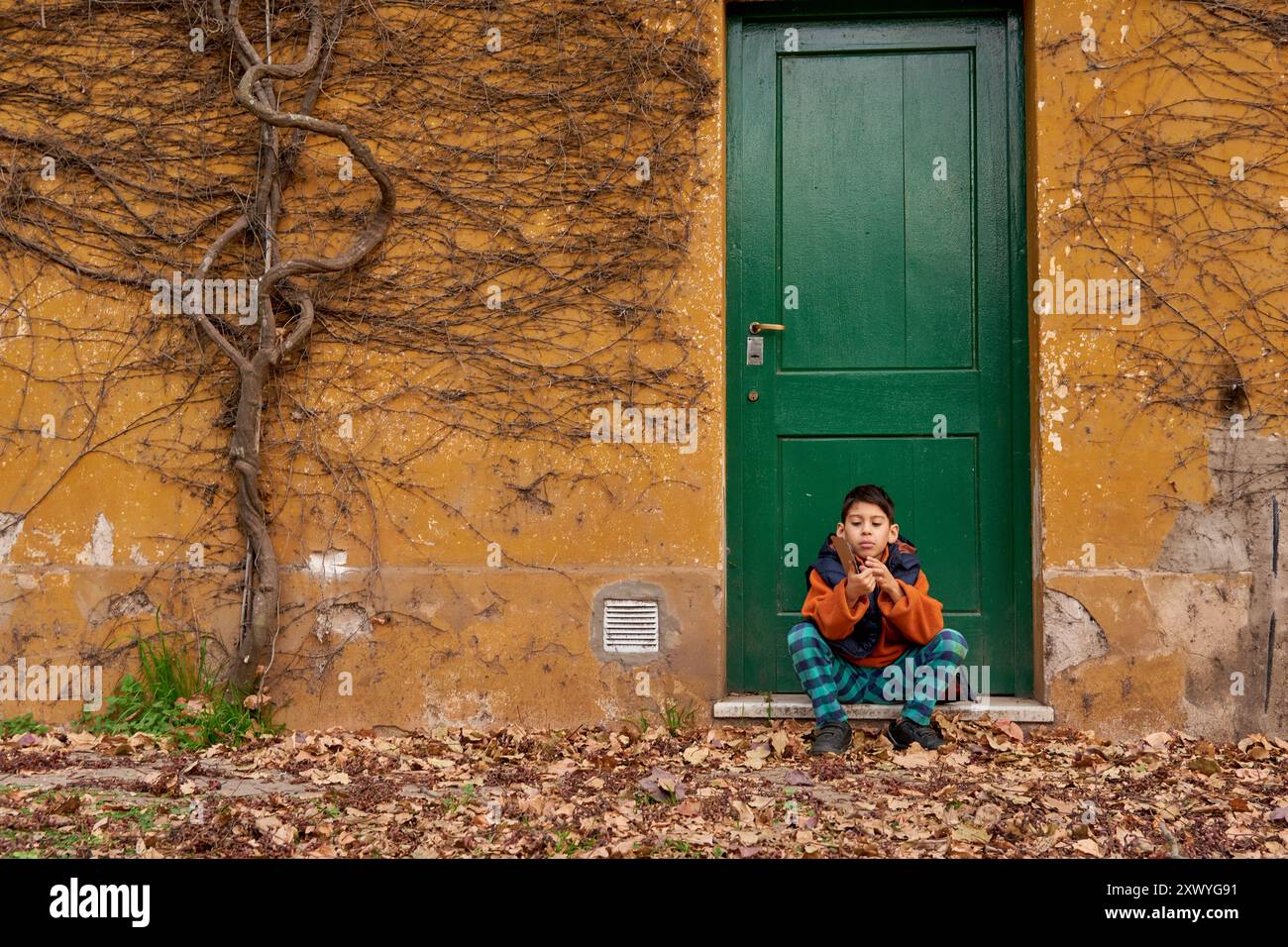 ragazzo latino seduto di fronte ad una casa arancione con una porta verde con una vite secca attaccata al muro e foglie sul pavimento in un pomeriggio autunnale. Foto Stock