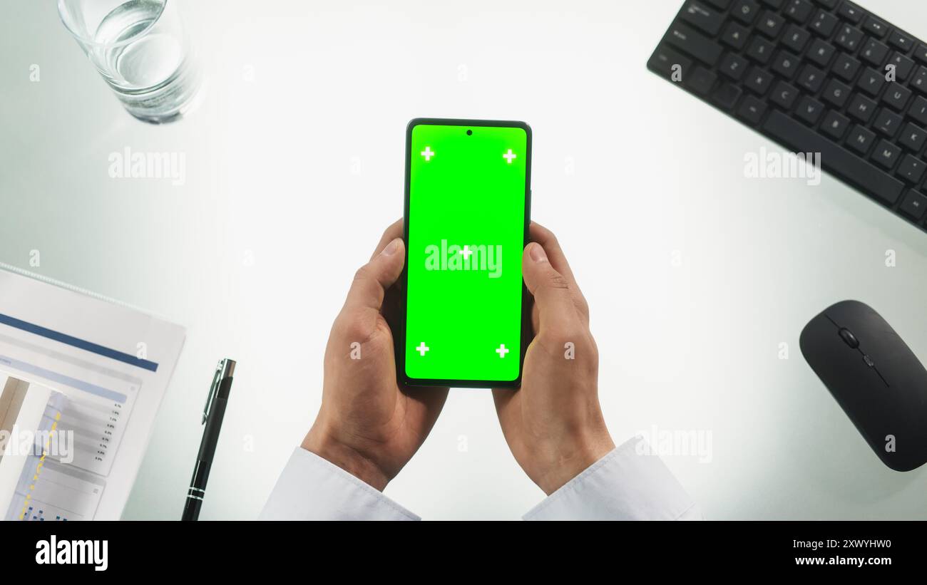 Persona anonima che utilizza uno smartphone con schermo Chromakey con occultamento verde e segnaposto. Uomo che tiene il cellulare verticalmente con entrambe le mani, strisciando e toccando il telefono. Vista dall'alto verso il basso Foto Stock