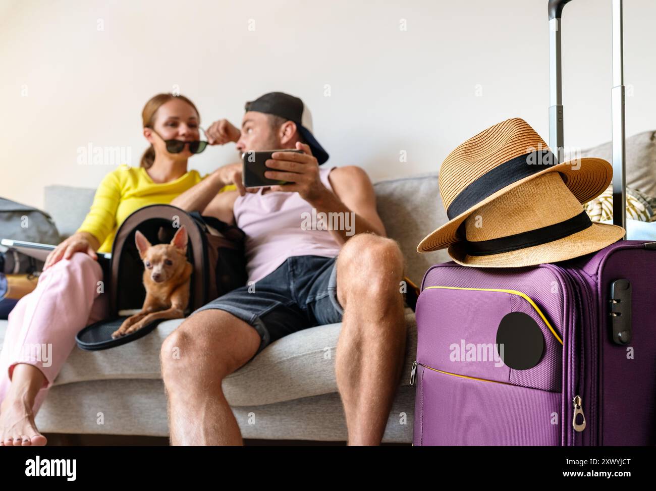 Borsa da viaggio con cappelli di paglia su sfondo sfocato, con una coppia felice con un cane piccolo che prenota il loro viaggio estivo. Foto Stock