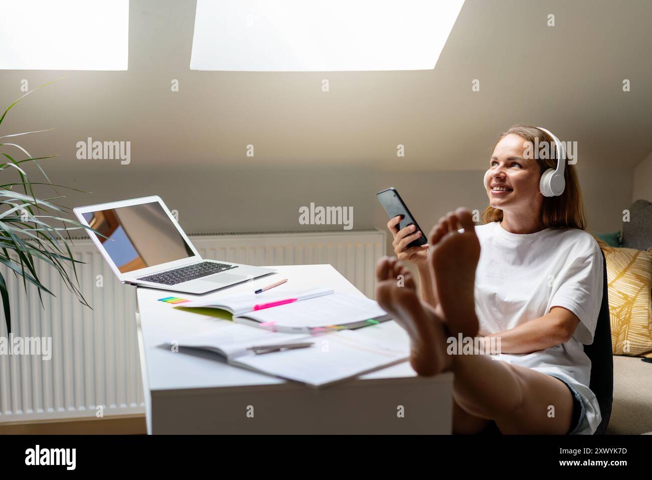 Giovane studentessa che indossa t-shirt bianca, donna che studia a una scrivania a casa. Ascolta il podcast educativo. Foto Stock