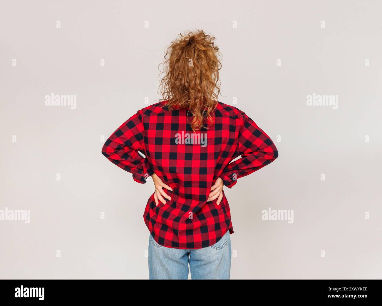 Vista posteriore della donna che indossa una camicia a quadri rossa con le mani in vita isolata su sfondo grigio chiaro. Foto Stock