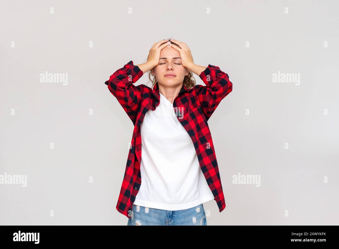 Donna caucasica che indossa una camicia casual rossa a quadri si presenta su sfondo grigio chiaro e tiene la testa con le mani. Foto Stock