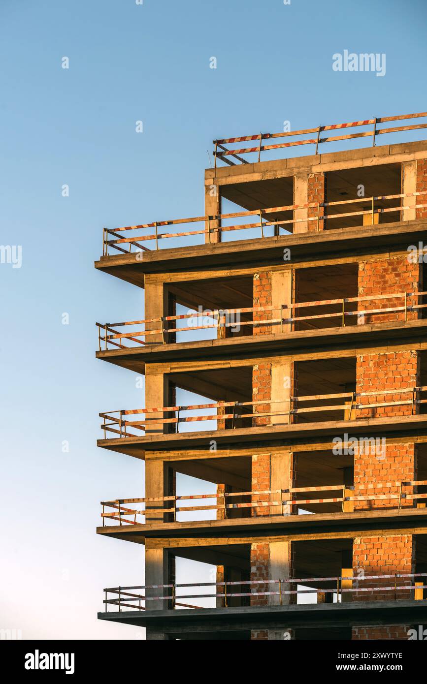Un edificio parzialmente costruito mostra mattoni grezzi e cemento con barriere di sicurezza in posizione durante l'ora d'oro serale. Foto Stock