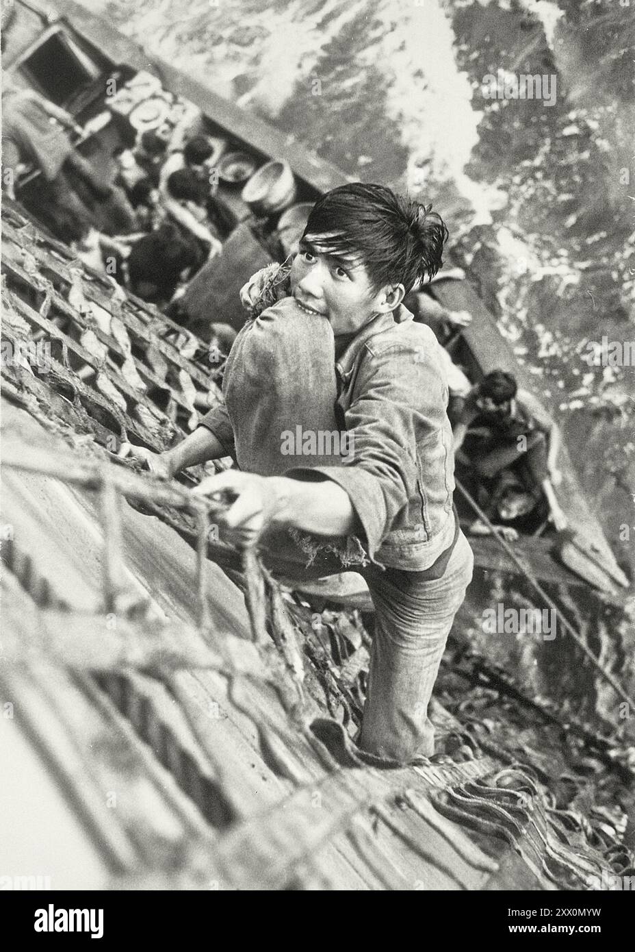 Guerra del Vietnam. Mar Cinese meridionale... Un rifugiato vietnamita con i suoi effetti personali fissati tra i denti, sale una rete di carico al ponte della nave da combattimento USS White Plains, AFS-M. La nave sta raccogliendo ventinove rifugiati da una barca di legno di 35 piedi. anni '1970 Foto Stock