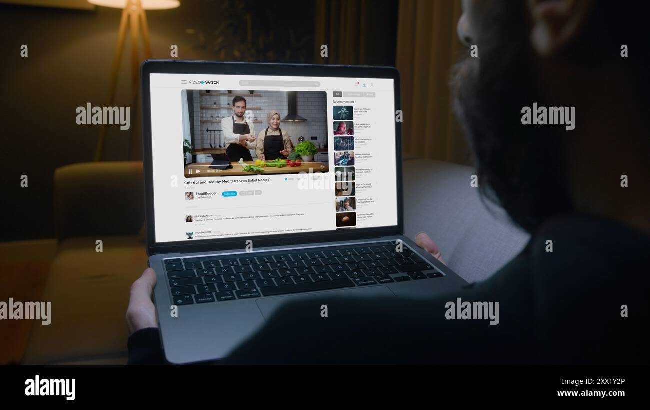Uomo sdraiato sul divano che guarda il video di una coppia di food blogger su un portatile, che impara a preparare una sana insalata mediterranea in salotto di notte. Foto Stock