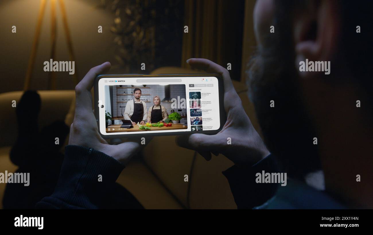 Uomo sdraiato sul divano che guarda un tutorial di cucina sul suo smartphone in salotto. L'apprendimento digitale moderno e la comodità di accedere all'istruzione Foto Stock