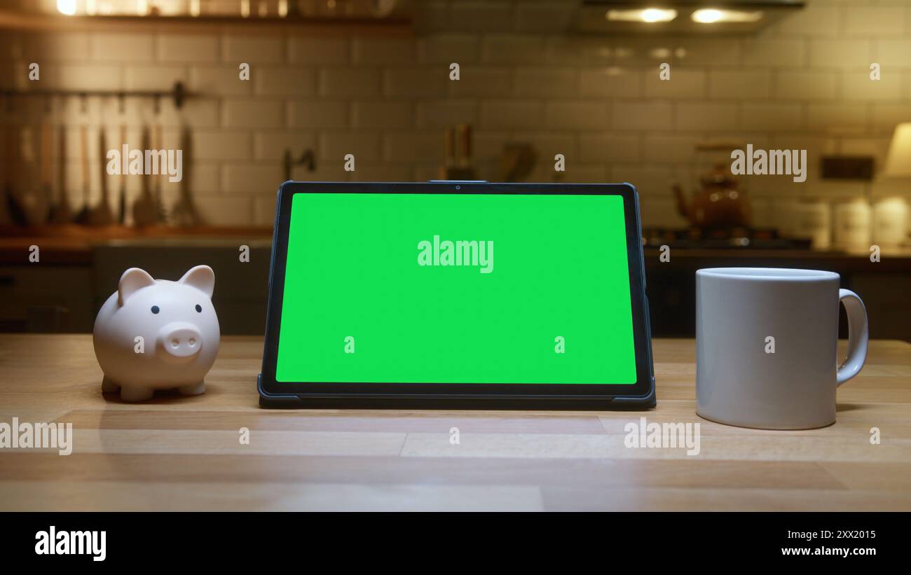 Un tablet digitale con schermo verde su un bancone della cucina con salvadanaio e una tazza da caffè bianca in una cucina accogliente. Foto Stock