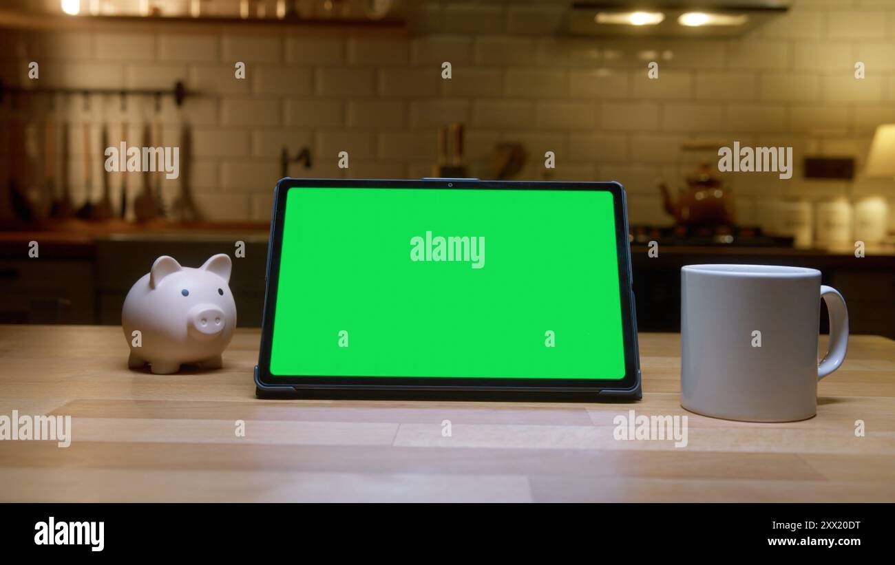 Un tablet con schermo verde su un tavolo di legno, con tazza bianca e un salvadanaio. Gestione finanziaria e tecnologia in un'accogliente cucina casalinga Foto Stock