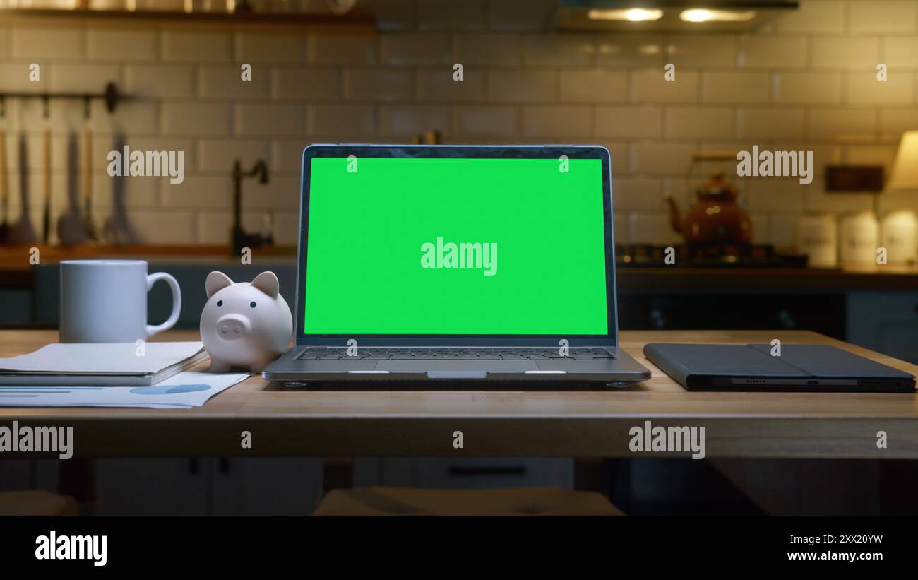 Un notebook con schermo verde su un bancone della cucina con una tazza da caffè bianca e un salvadanaio accanto in una cucina moderna. Foto Stock