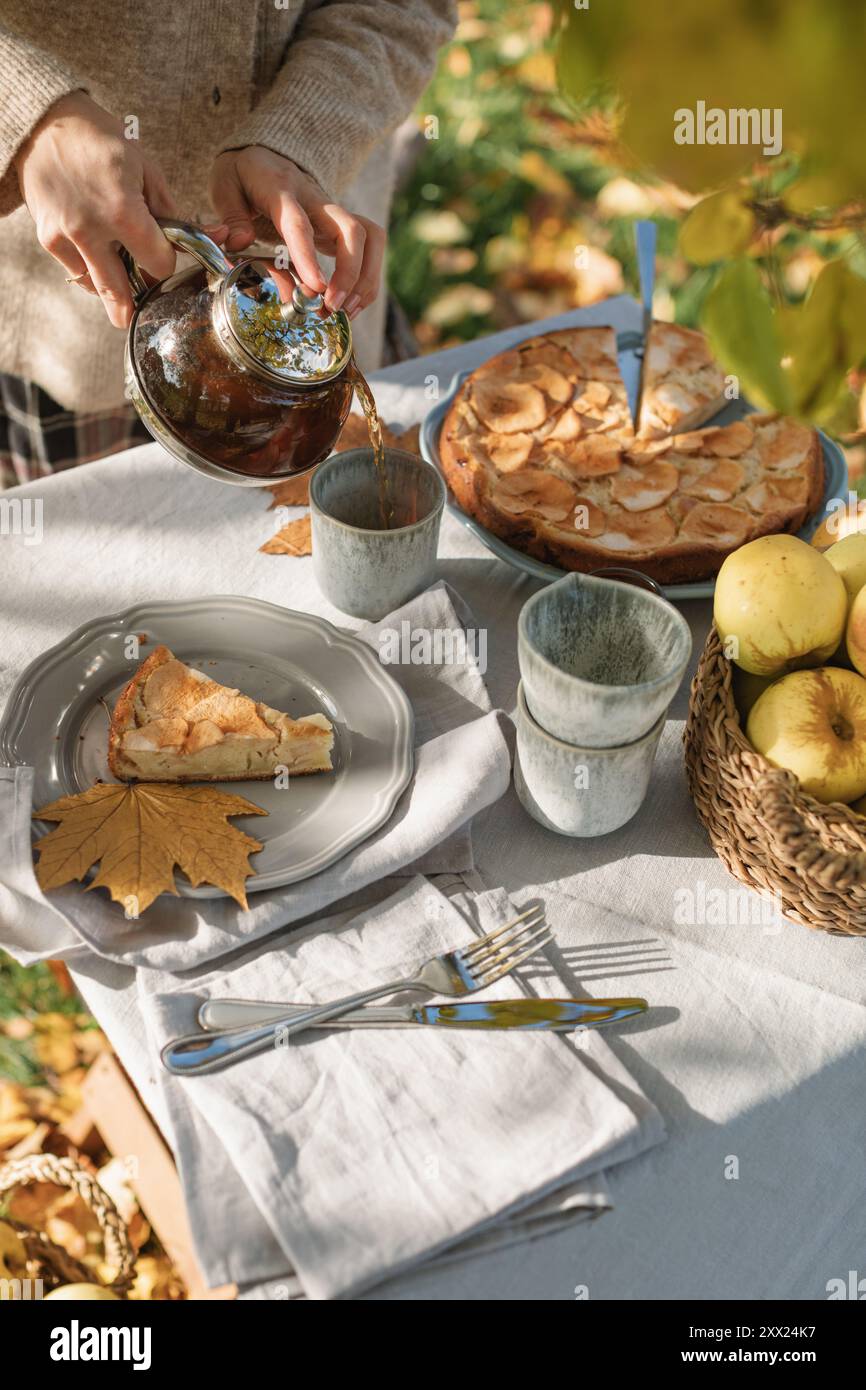 Giovane donna che versa una tazza di tè e un tavolo con una torta di mele e mele fresche su un tavolo da giardino autunnale al sole Foto Stock