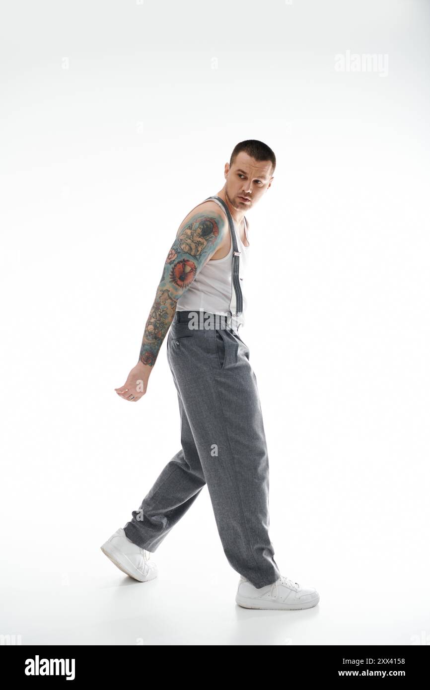 Un uomo con un tatuaggio a manica intera cammina con sicurezza in una canotta bianca e pantaloni grigi. Foto Stock